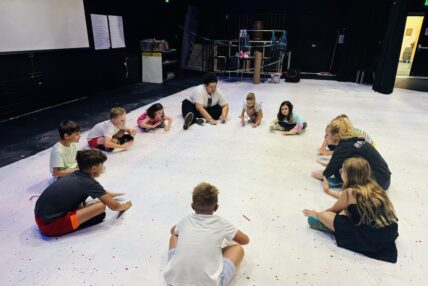 Two teachers leading a group of students in drumming on the floor.