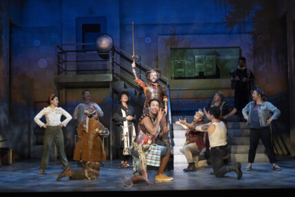 A man stands on stage holding a sword upward as the cast surrounds him, kneeling.