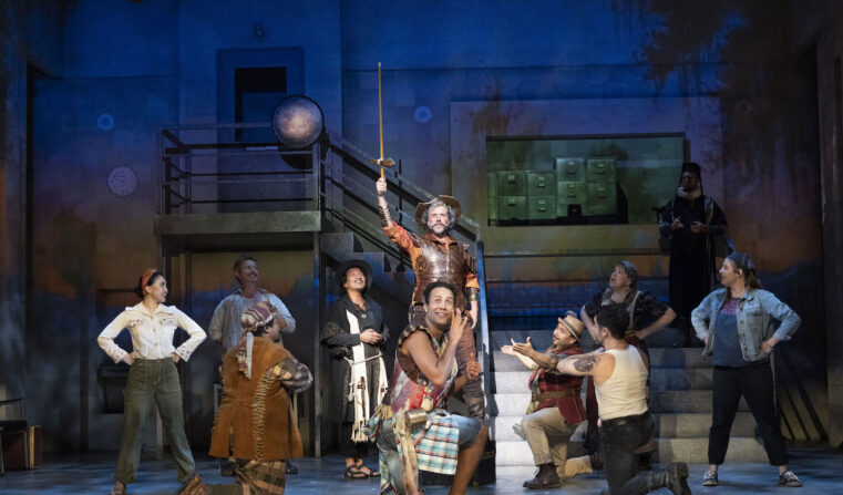 A man stands on stage holding a sword upward as the cast surrounds him, kneeling.