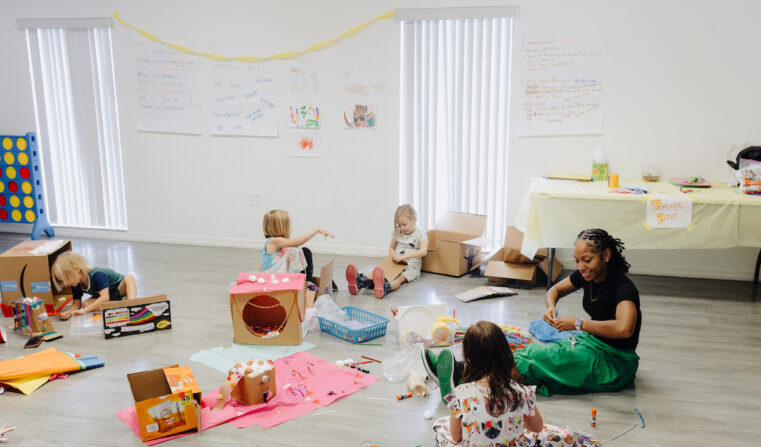 A female teacher surrounded by kids in a classroom doing arts and crafts.