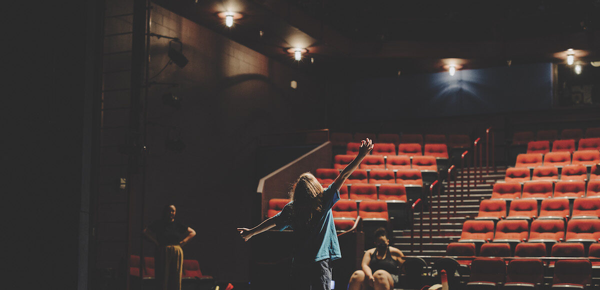 Shot from behind, a young actor stands with outstretched arms in front of empty auditorium seats as two people watch.