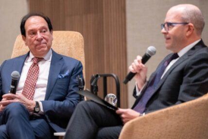 Two men in suits speak into microphones for an audience.