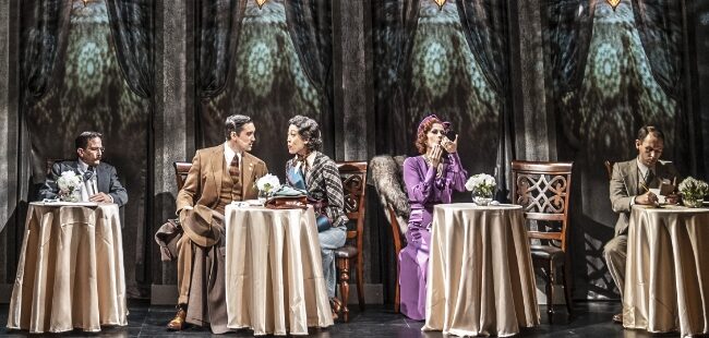 Actors in period attire sit at small tables on stage.