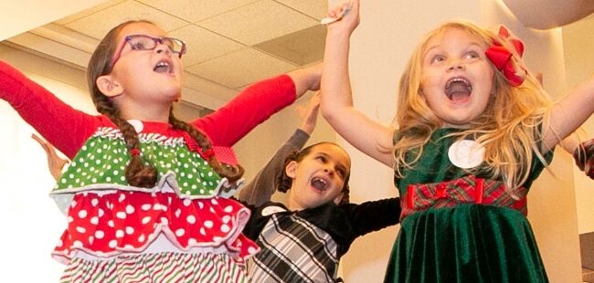 Three young girls in holiday attire holding their arms up and smiling.