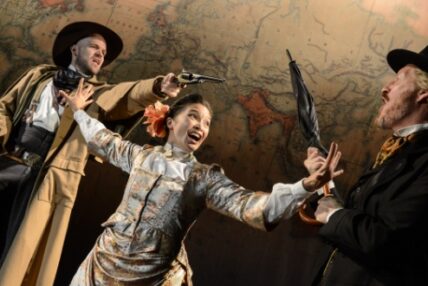 A woman stands between two men pointing prop guns at one another.