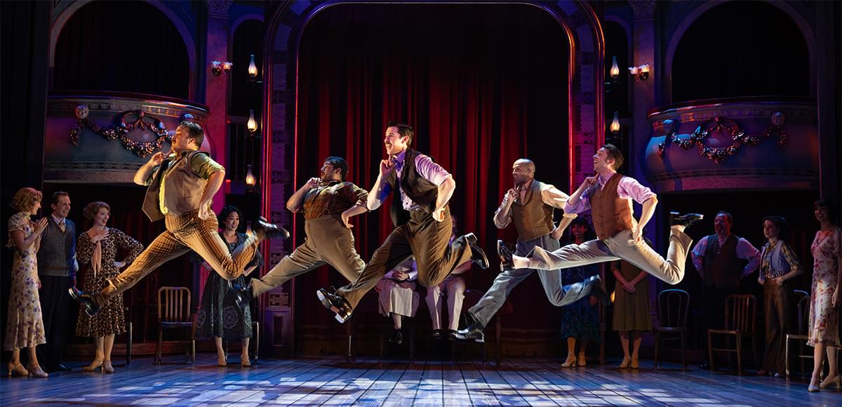 Asolo stage performance with male dancers in mid-jump and acting spectators watching.