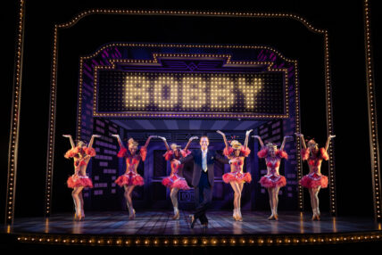 A man in a suit in a presentation stance with six female cabaret-style dancers behind him.