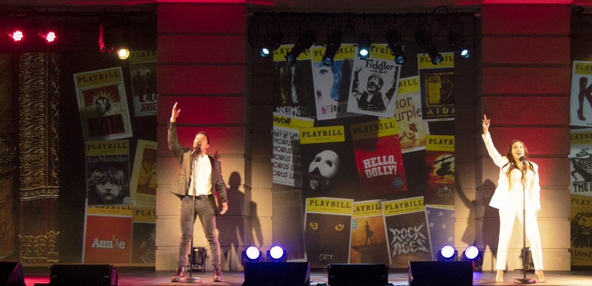 Two performers singing on stage against a backdrop of playbills.