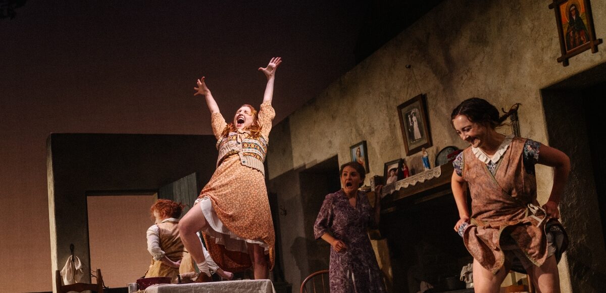 A woman sings on top of a table while other women smile, dance and clean.