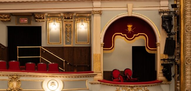 An empty balcony showing white or light gray walls with gold trim and filigree and red fabric seats and banister tops.