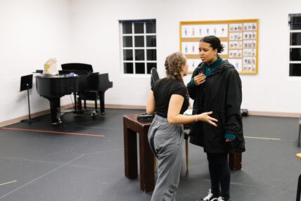 Two women singing in front of a piano during rehearsal.