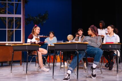 A group of men and women sitting at school desks on stage and talking.