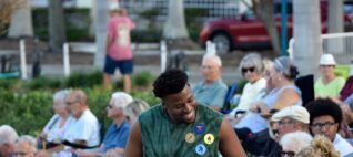 A young Black man stands chatting to another audience member at an outdoor performance.