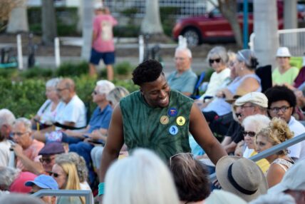 A young Black man stands chatting to another audience member at an outdoor performance.