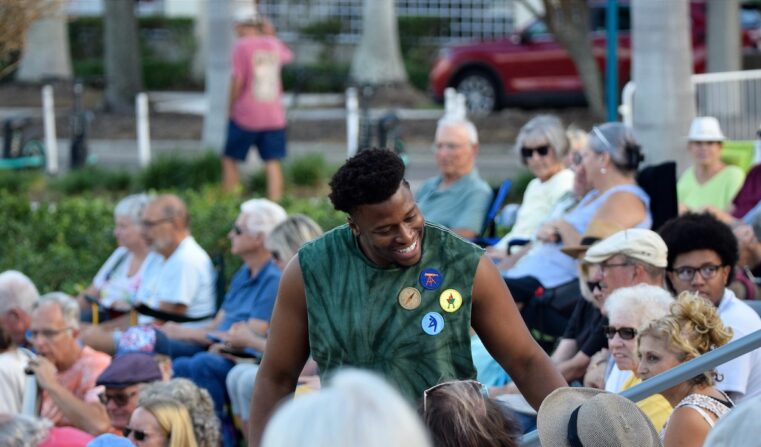 A young Black man stands chatting to another audience member at an outdoor performance.