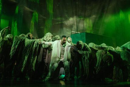 An actor in a white shroud surrounded by cast members in hooded gray shrouds.