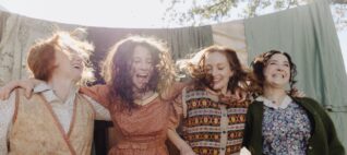 four women in costumes depicting the working class of the early 1900s all are standing side by side smilling in front of a clothesline