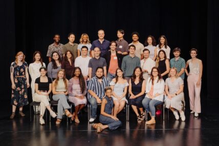 A diverse group of about thirty students in three rows (two standing one sitting) and one person seated on the floor in front of them.