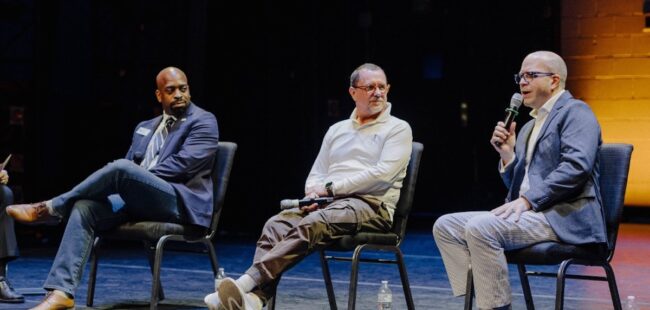 Three men sit on stage, speaking to the audience.