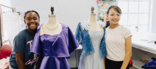 in the theatre costume shop, two yong women smiling, standing on either side of a pair of manaquins each with a costume dress