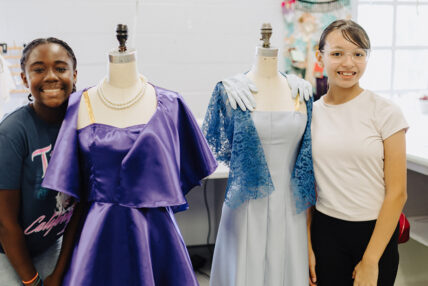 in the theatre costume shop, two yong women smiling, standing on either side of a pair of manaquins each with a costume dress