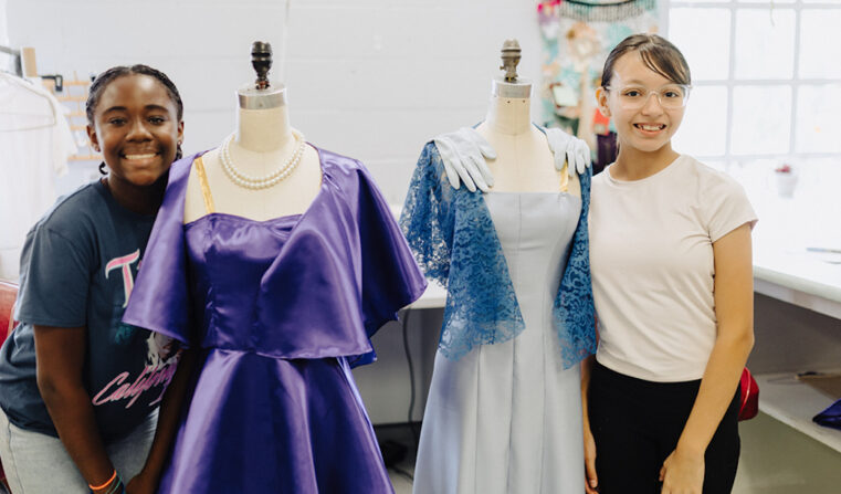 in the theatre costume shop, two yong women smiling, standing on either side of a pair of manaquins each with a costume dress