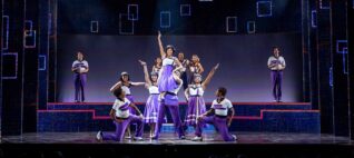 twelve diverse performers at the end of a dance routine with a man holding up an actress and the pair surrounded by others peformers in various stances all are wearing purple and white costumes reminiscent of 1950 high school or college gym class