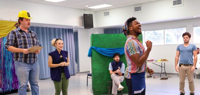 A man singing during rehearsal while his cast-mates look on.