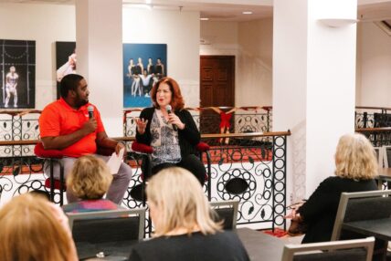 A man and woman holding microphones talking before a small audience.