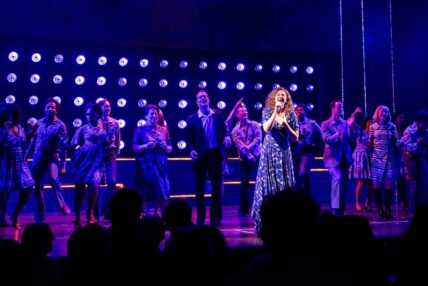 A woman in the spotlight singing while surrounded by a large group of men and women signing and dancing.
