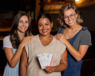 Three female patrons standing in close proximity smiling the center one holds three tickets.