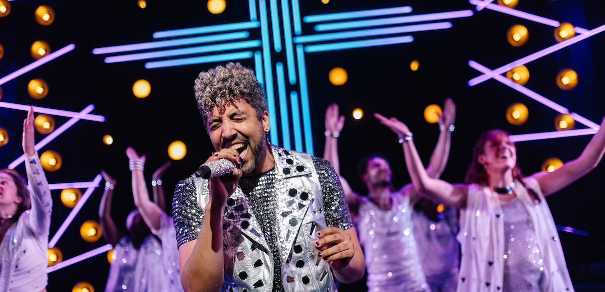 A Black male singing while six dancers perform behind him.