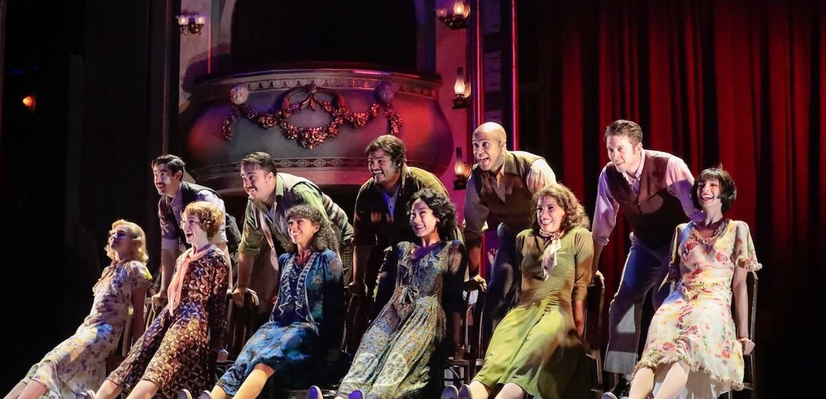 Six seated actresses outstretching their legs while five actors lean on the chair backs.