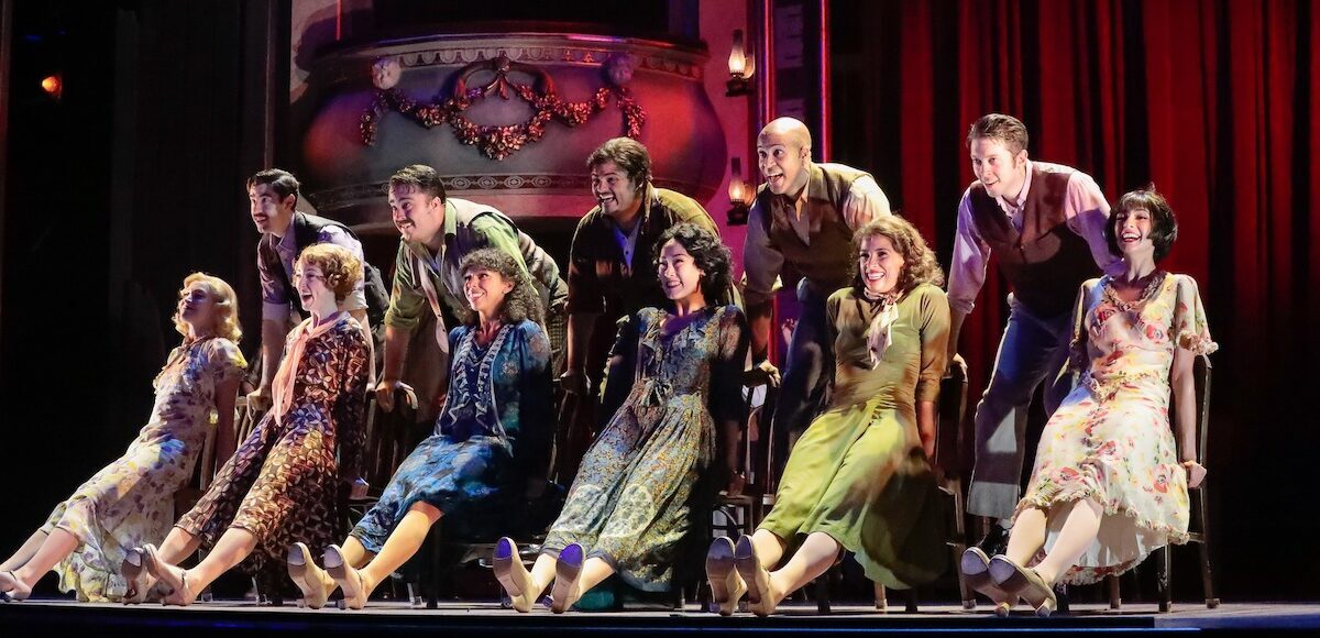 Six seated actresses outstretching their legs while five actors lean on the chair backs.
