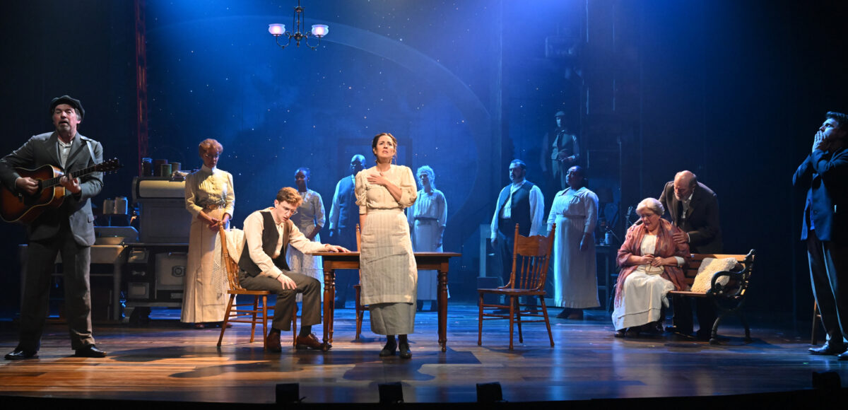 Actor and actress sit a table at centerstage as the ensemble cast looks on.