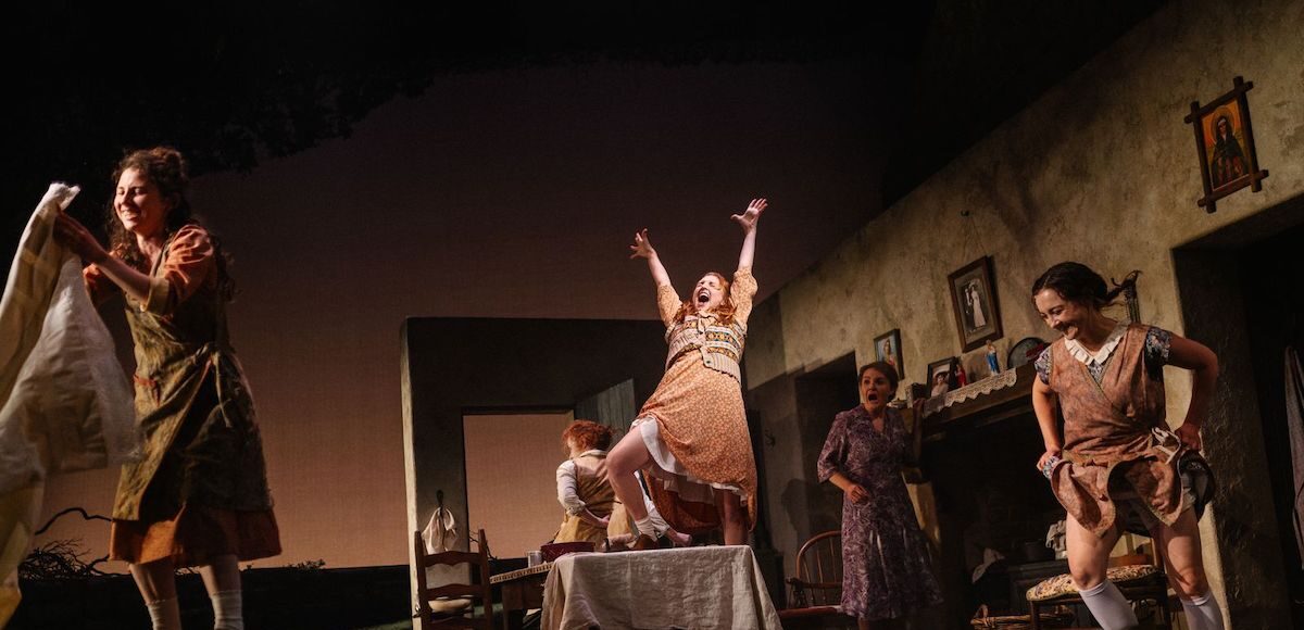 A woman sings on top of a table while other women smile, dance and clean.