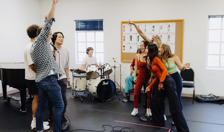 A group of men and a group of women sing at each other while a man plays the drums in the background.