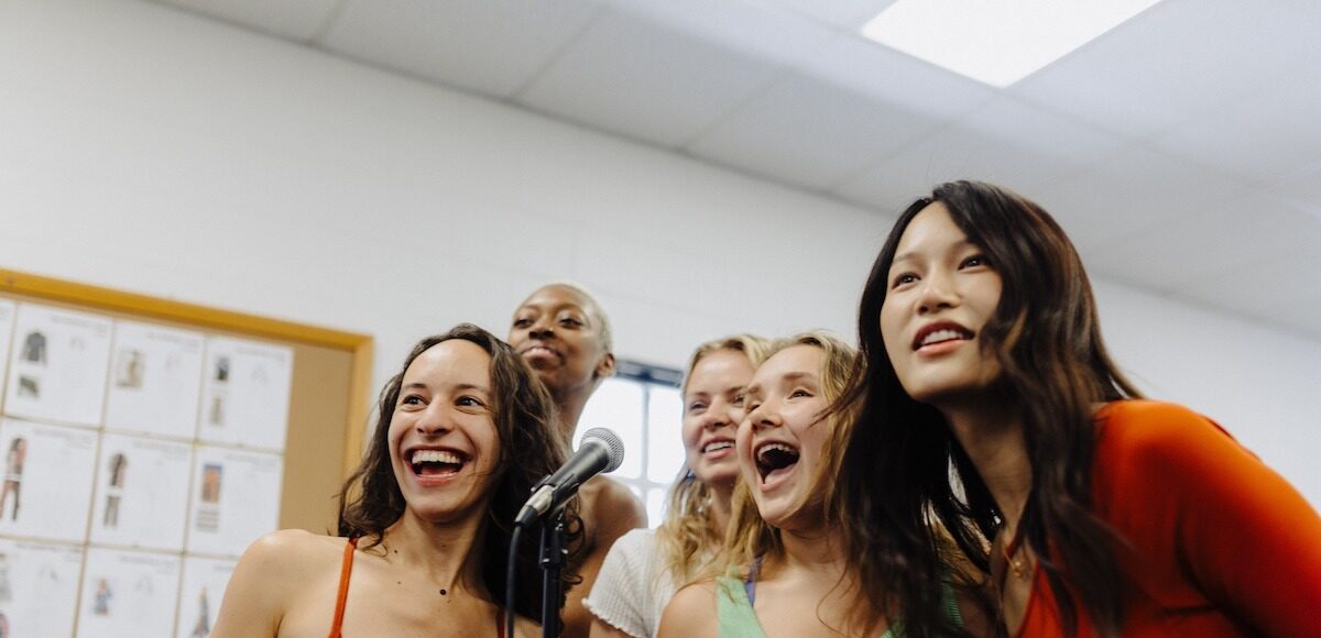 Five women singing into a standing microphone in a rehearsal room