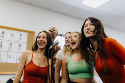 Five women singing into a standing microphone in a rehearsal room