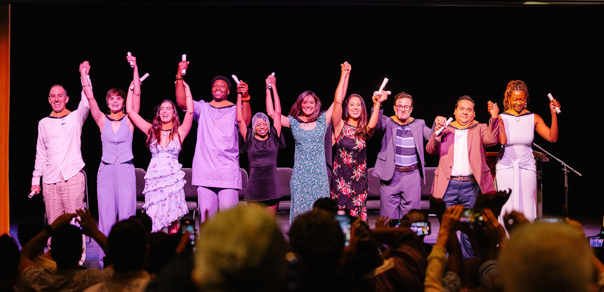 A line of people holding hands and rolled up papers in the air before an audience.