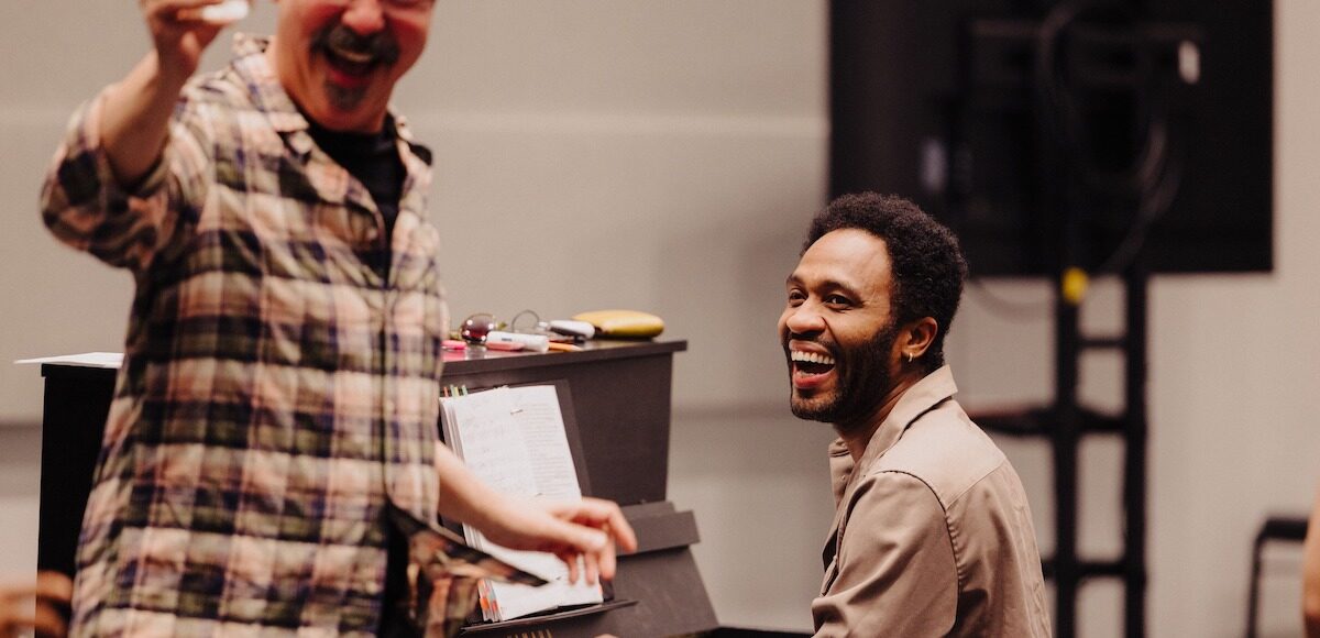 A man plays the piano, smiling and laughing as another man sings.