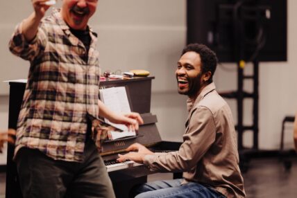A man plays the piano, smiling and laughing as another man sings.