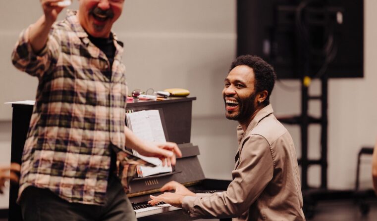 A man plays the piano, smiling and laughing as another man sings.