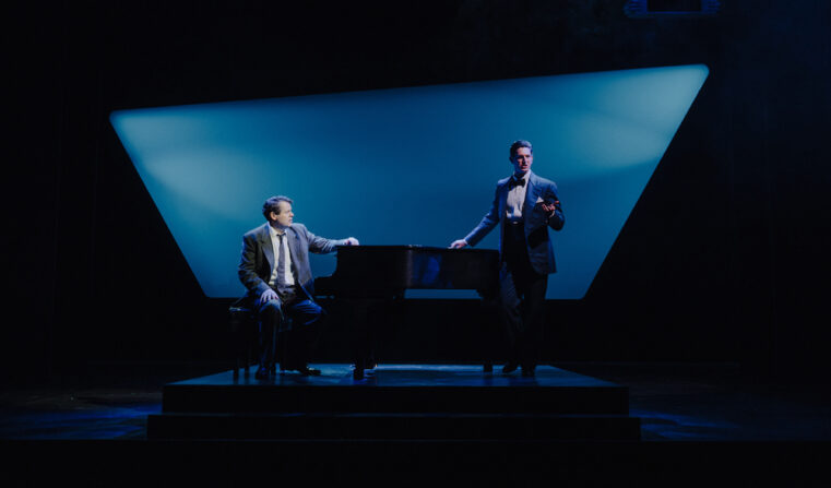 Two men on a darkened stage one seated at a piano and the other standing on the other side of the piano.