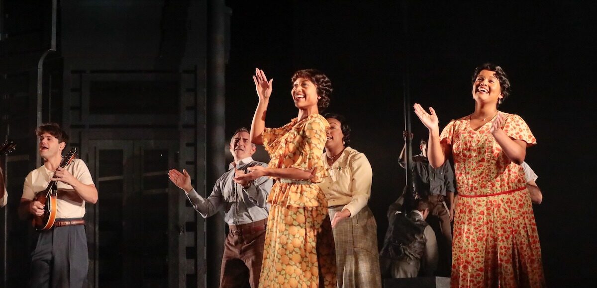 A group of men and women in period attire sing, dance and clap.
