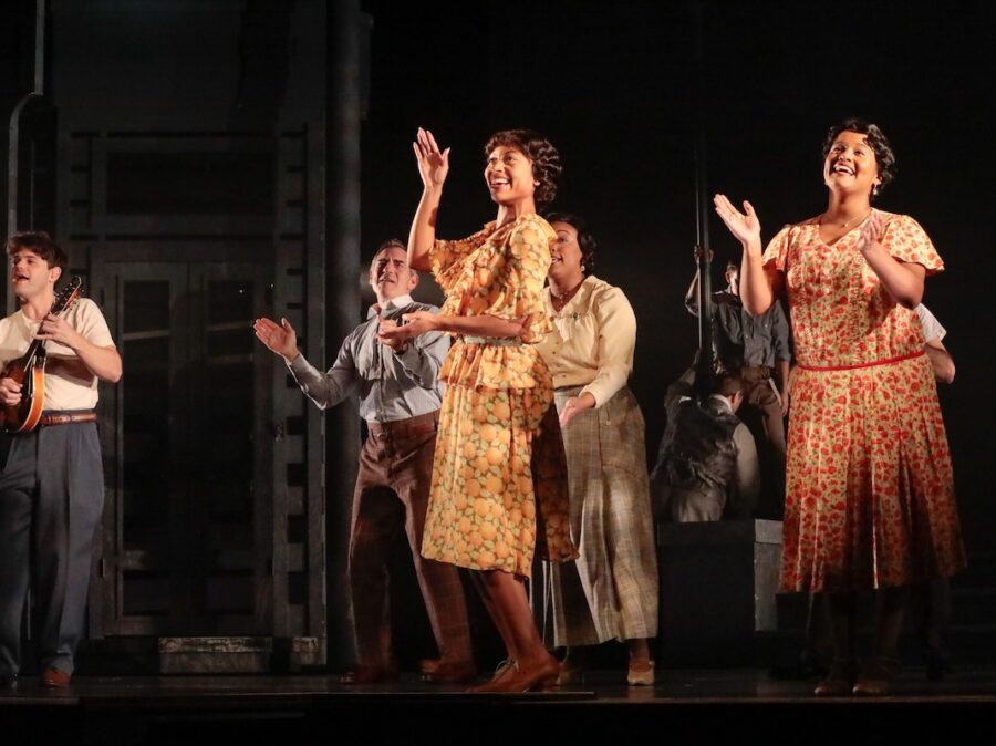 A group of men and women in period attire sing, dance and clap.