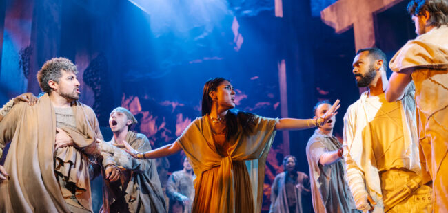 An actress mid-performance surrounded by costumed male actors.