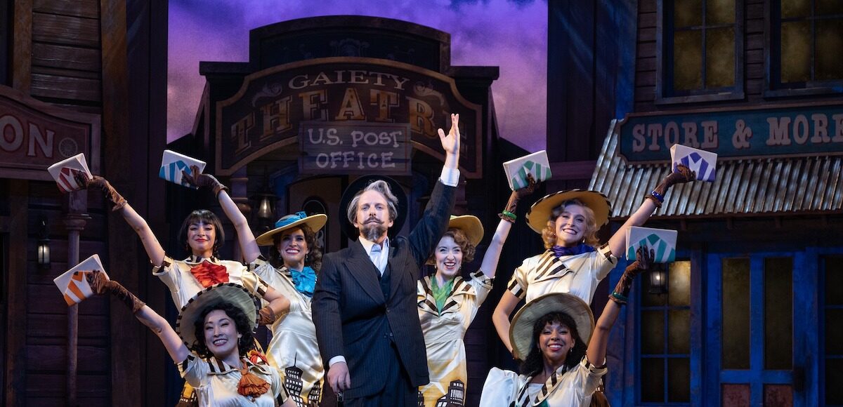 An actor with a fancy mustache poses between six women in front of a historic town set.