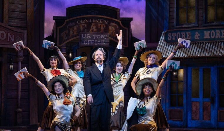 An actor with a fancy mustache poses between six women in front of a historic town set.