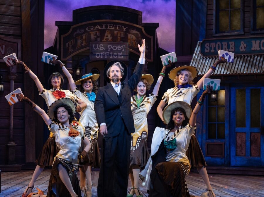 An actor with a fancy mustache poses between six women in front of a historic town set.